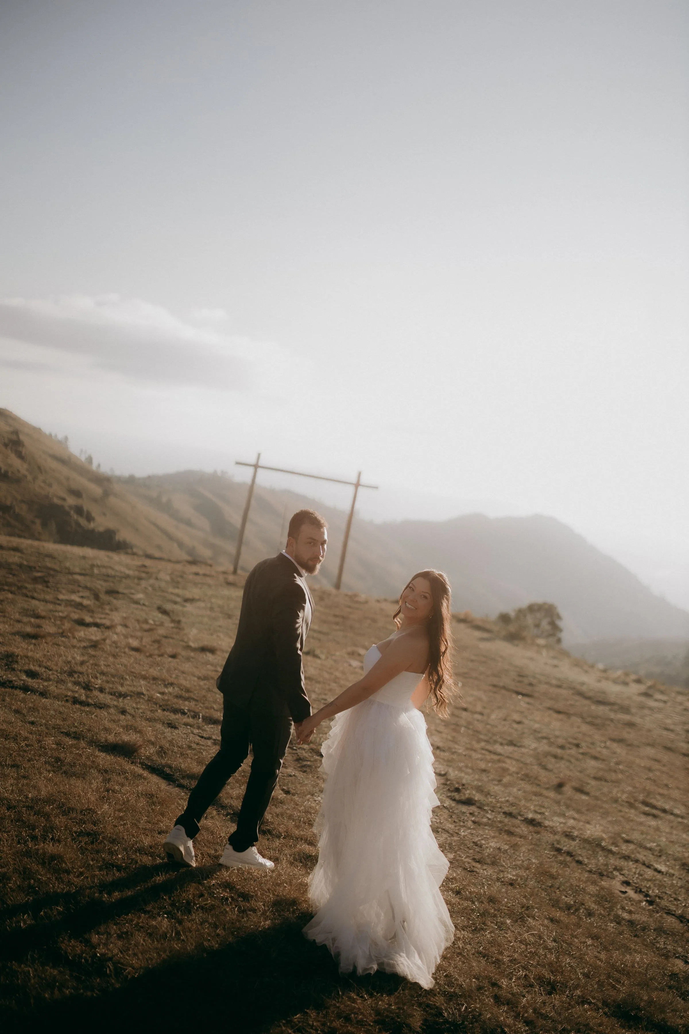 The couple walk hand in hand across a hillside, a simple wooden arch behind them