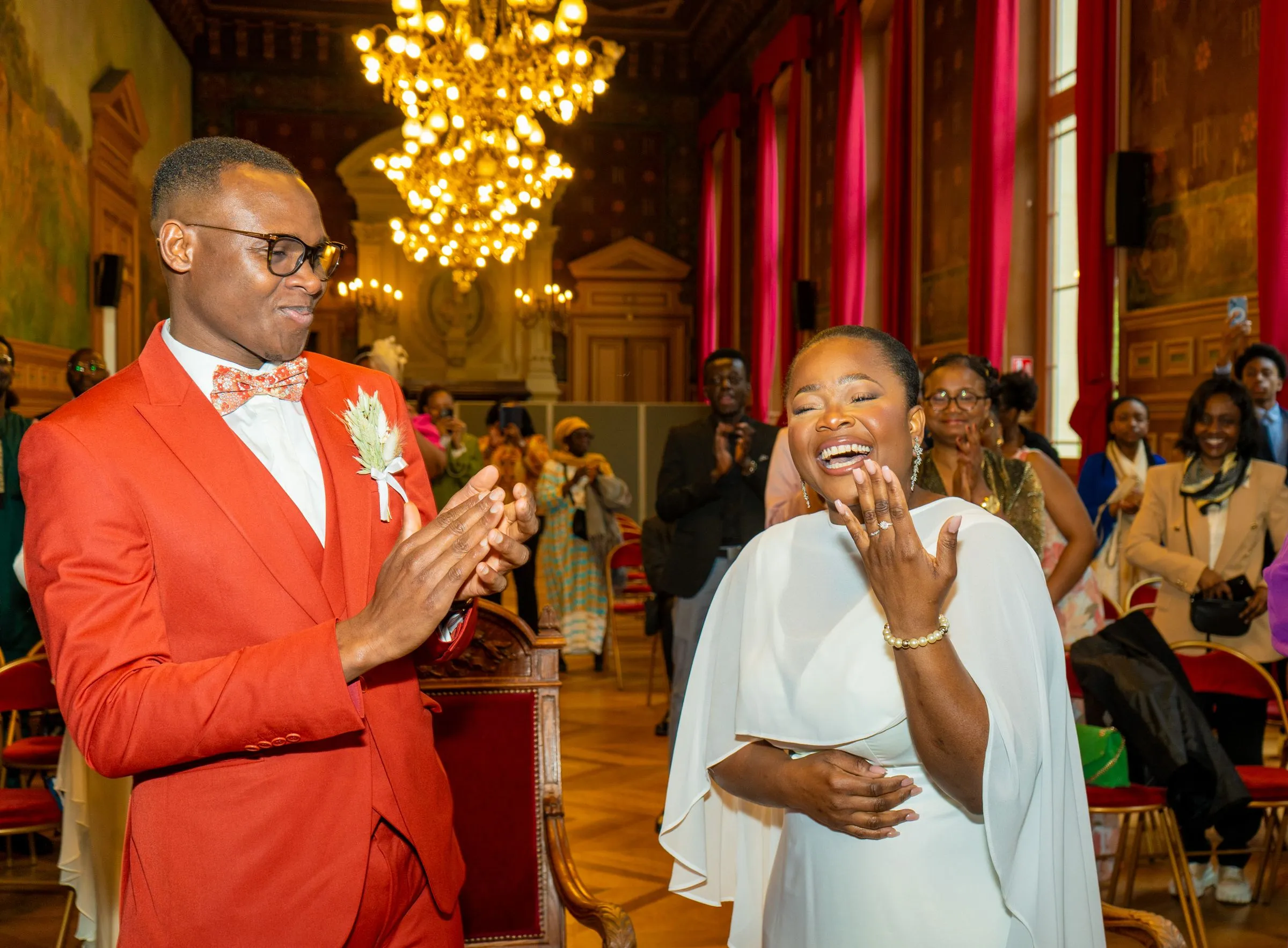 The couple clap and laugh as they walk into a grand hall with red curtains and a chandelier